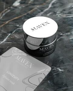Round white MavenX Longevity cream jar on a dark marble surface with a reflective tin beside it, suggesting luxury skincare packaging