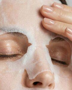 Person smooths a white facial sheet mask onto their face, focusing around the cheeks and nose.