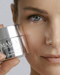 Close-up of a woman's face holding a silver jar labeled 'MAVEX Switzerland Longevity Supreme Cream' against a light background, showcasing the product near the eye.