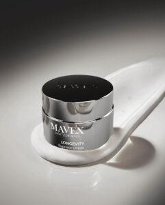 Close-up of a shiny silver MAVTEX Longevity Supreme Cream jar on a white curved display surface with a dark-to-light gradient background.
