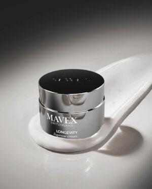 Close-up of a shiny silver MAVTEX Longevity Supreme Cream jar on a white curved display surface with a dark-to-light gradient background.