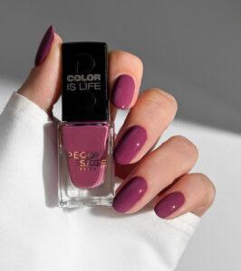 Hand holding a pink-purple nail polish bottle labeled Color Is Life with matching painted nails on a white sleeve backdrop.
