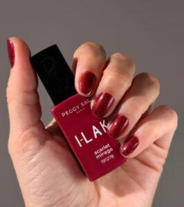 Close-up of a hand holding a Peggy Sage I-LAK red nail polish bottle with matching glittery red nails visible on all fingers.