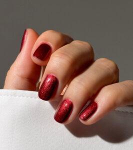 Close-up of a hand showing glittery red manicure on short oval nails, resting on white fabric.