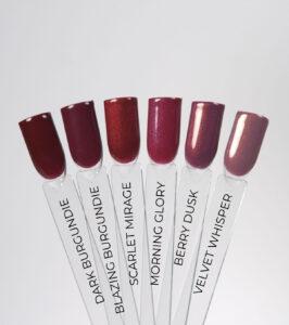 Swatches of six wine and burgundy nail polishes labeled from left: Dark Burgundy, Blazing Burgundy, Scarlet Mirage, Morning Glory, Berry Dusky, Velvet Whisperer (nail tips on clear sticks).