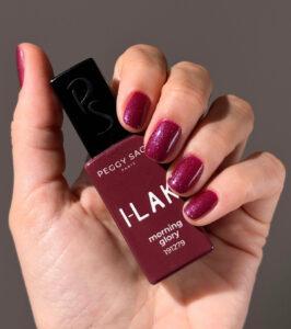 Close-up of a hand holding Peggy Sage I-LAK Morning Glory nail polish in deep magenta with shimmer, nails painted to match the bottle's color.