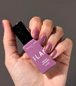 Close-up of a hand with lavender-pink manicured nails holding a Peggy Sage I-LAK Velvet Whisper nail polish bottle.